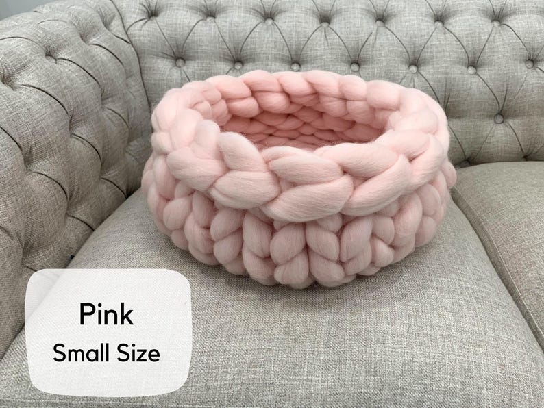 May include: A small, round, light pink basket crafted from thick, chunky knit yarn. The basket features a braided, interlocking texture throughout its circular structure. A white label in the corner displays the text Pink Small Size.
