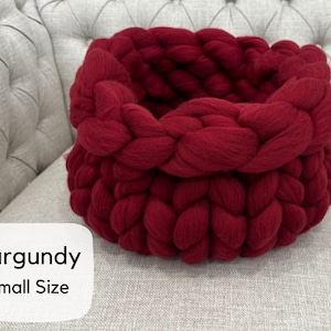 May include: A round, deep basket crafted from thick, chunky burgundy yarn in a braided knit pattern. The basket features a wide, folded-over rim. A white label in the bottom left corner displays the text Burgundy Small Size in black font.
