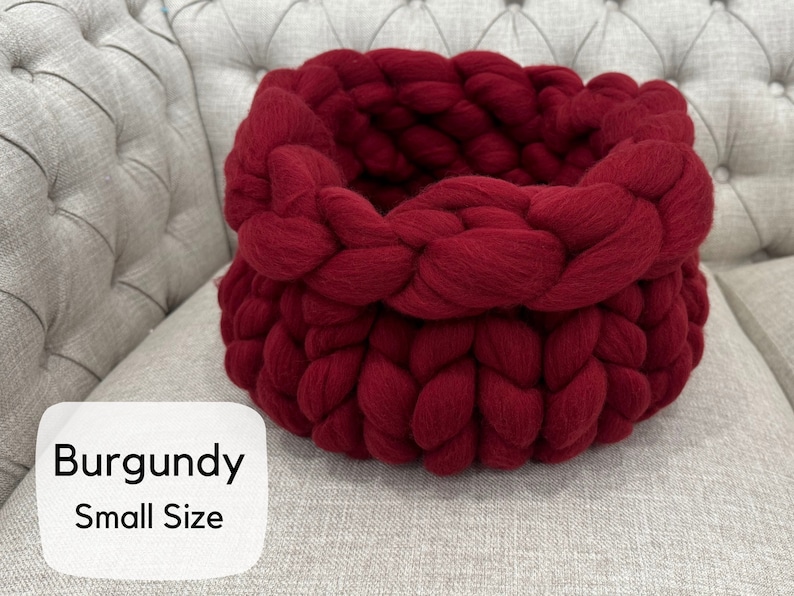 May include: A round, deep basket crafted from thick, chunky burgundy yarn in a braided knit pattern. The basket features a wide, folded-over rim. A white label in the bottom left corner displays the text Burgundy Small Size in black font.