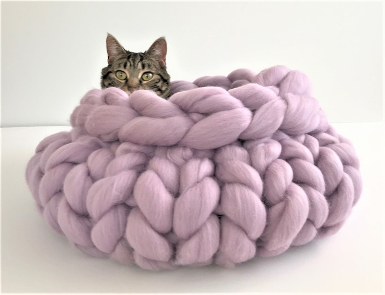 May include: A round, chunky knit pet bed made of thick, soft lavender yarn with a braided texture. The bed features a deep, circular basin with high, plush walls. A tabby cat with green eyes is partially visible, resting inside the lavender bed.