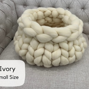 May include: A round, chunky knit basket crafted from thick, ivory-colored yarn. The basket features a prominent braided texture throughout its structure. A white label in the bottom left corner displays the text Ivory Small Size in black font.