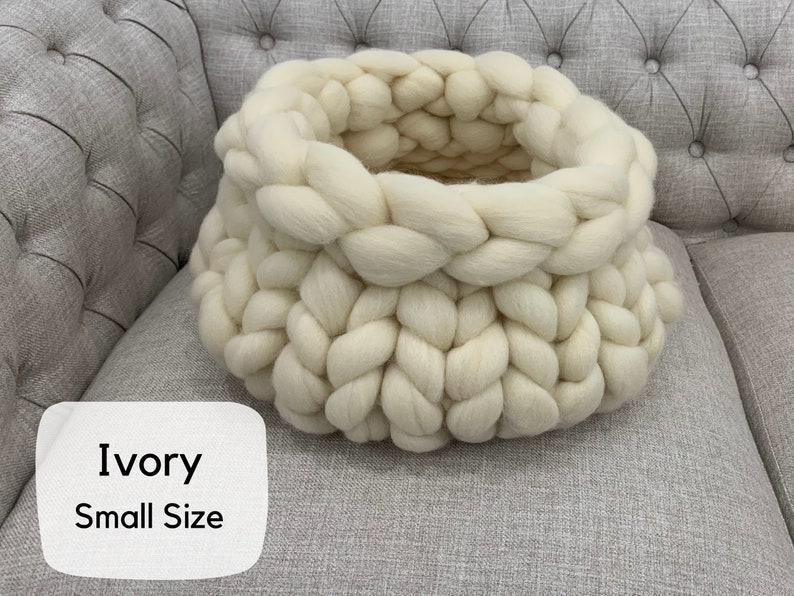 May include: A round, chunky knit basket crafted from thick, ivory-colored yarn. The basket features a prominent braided texture throughout its structure. A white label in the bottom left corner displays the text Ivory Small Size in black font.