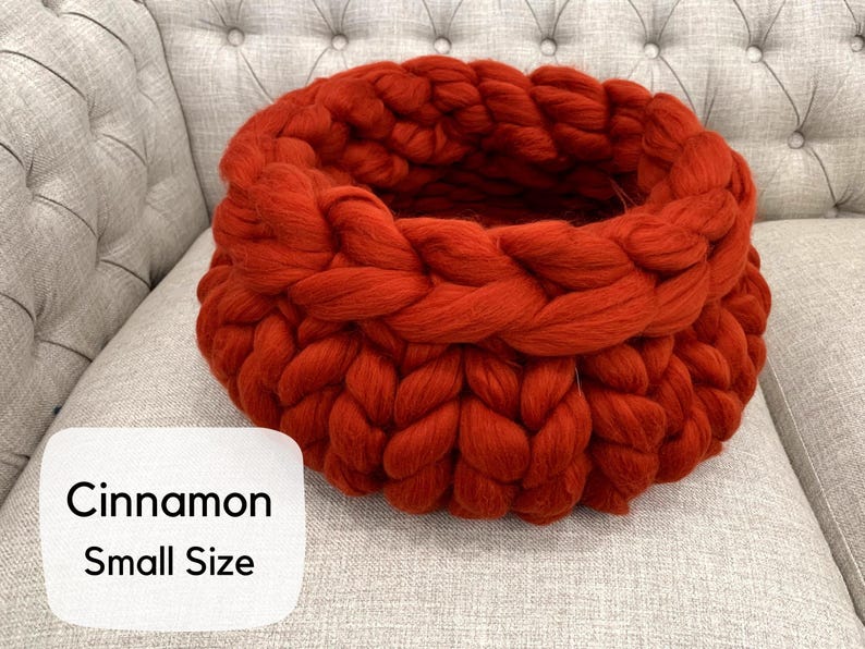 May include: A round, chunky knit basket crafted from thick, rust-orange yarn with a prominent braided texture. A white rectangular label in the lower left corner features the black text Cinnamon Small Size, indicating the color and size.