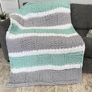 May include: A chunky knit blanket in a color block design. The blanket features wide stripes of mint green, white, and gray. The soft, textured blanket is draped over a gray sofa. This cozy throw blanket is perfect for snuggling.
