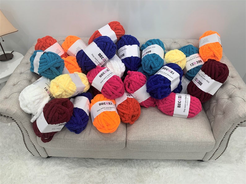 May include: A variety of colorful yarn skeins in different shades of red, orange, yellow, green, blue, purple, and white. The yarn is arranged on a light gray couch. The yarn is labeled "Beczi" and says "Stay warm and Be Cozy".