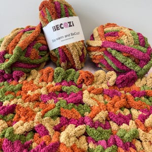 Peut inclure: Trois pelotes de laine dans des tons de vert, orange, rose et beige. La laine est de texture épaisse et duveteuse. La laine est étiquetée "BeCozi Stay warm and BeCozi! www.becozi.net"