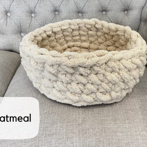 Puede incluir: Una cesta redonda de punto grueso color avena. La cesta est&aacute; hecha de un hilo suave y texturizado y se encuentra sobre una superficie tapizada gris. La palabra "Oatmeal" se muestra en una etiqueta blanca.