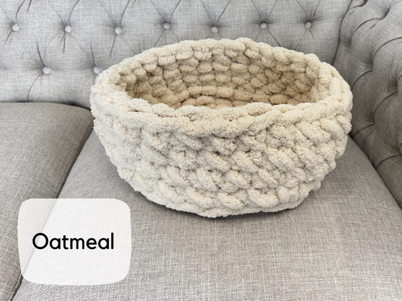 Puede incluir: Una cesta redonda de punto grueso color avena. La cesta est&aacute; hecha de un hilo suave y texturizado y se encuentra sobre una superficie tapizada gris. La palabra "Oatmeal" se muestra en una etiqueta blanca.