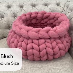 May include: A round, chunky knit basket crafted from thick, blush pink yarn. The basket features a prominent braided texture throughout its circular structure. A white label on the lower left corner displays the text Blush Medium Size.