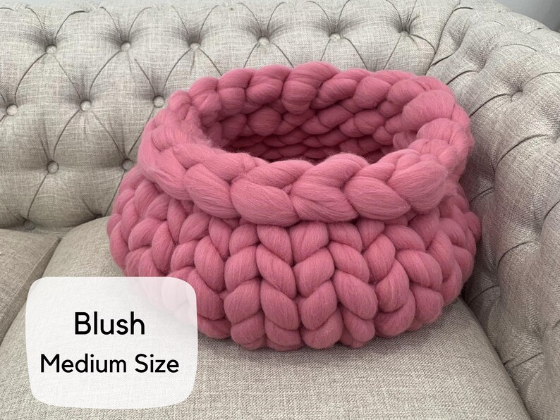 May include: A round, chunky knit basket crafted from thick, blush pink yarn. The basket features a prominent braided texture throughout its circular structure. A white label on the lower left corner displays the text Blush Medium Size.