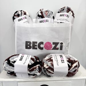Peut inclure: Cinq pelotes de laine dans un schéma de couleurs brun, noir, blanc et rose. La laine est dans un sac tote blanc avec le nom de la marque "BeCozi" imprimé sur le devant. La laine est étiquetée "Restez au chaud et BeCozi!"