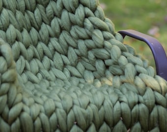 Puede incluir: Primer plano de una manta de punto grueso de color verde apagado. La manta está colocada sobre una silla de metal negro.