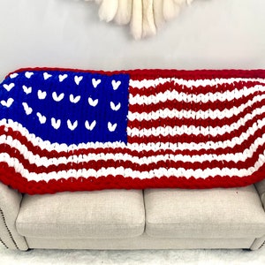 K&ouml;nnte beinhalten: Eine grob gestrickte Decke, die &uuml;ber einem Sofa drapiert ist und das Design der amerikanischen Flagge zeigt. Die Decke hat rote und wei&szlig;e Streifen, ein blaues Feld mit wei&szlig;en herzf&ouml;rmigen Sternen und einen roten Rand. Sie ist aus dickem, weichem Garn gefertigt.