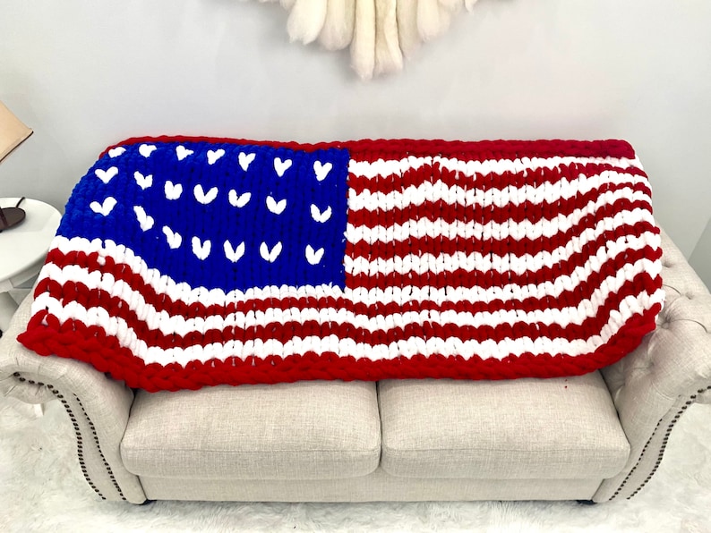K&ouml;nnte beinhalten: Eine grob gestrickte Decke, die &uuml;ber einem Sofa drapiert ist und das Design der amerikanischen Flagge zeigt. Die Decke hat rote und wei&szlig;e Streifen, ein blaues Feld mit wei&szlig;en herzf&ouml;rmigen Sternen und einen roten Rand. Sie ist aus dickem, weichem Garn gefertigt.