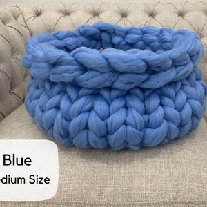May include: A round, chunky knit basket crafted from thick, soft blue yarn. The basket features a prominent braided texture throughout its circular structure. A white label in the bottom left corner displays the text "Blue" and "Medium Size".
