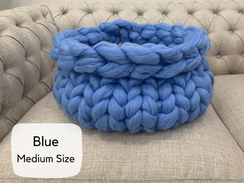 May include: A round, chunky knit basket crafted from thick, soft blue yarn. The basket features a prominent braided texture throughout its circular structure. A white label in the bottom left corner displays the text "Blue" and "Medium Size".