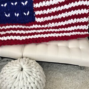 K&ouml;nnte beinhalten: Eine grob gestrickte Decke im Stil der amerikanischen Flagge mit roten und wei&szlig;en Streifen und einem blauen Feld mit wei&szlig;en Sternen. Ein wei&szlig;er, runder, gestrickter Pouf befindet sich im Vordergrund. Die Decke ist &uuml;ber ein wei&szlig;es Sofa drapiert.