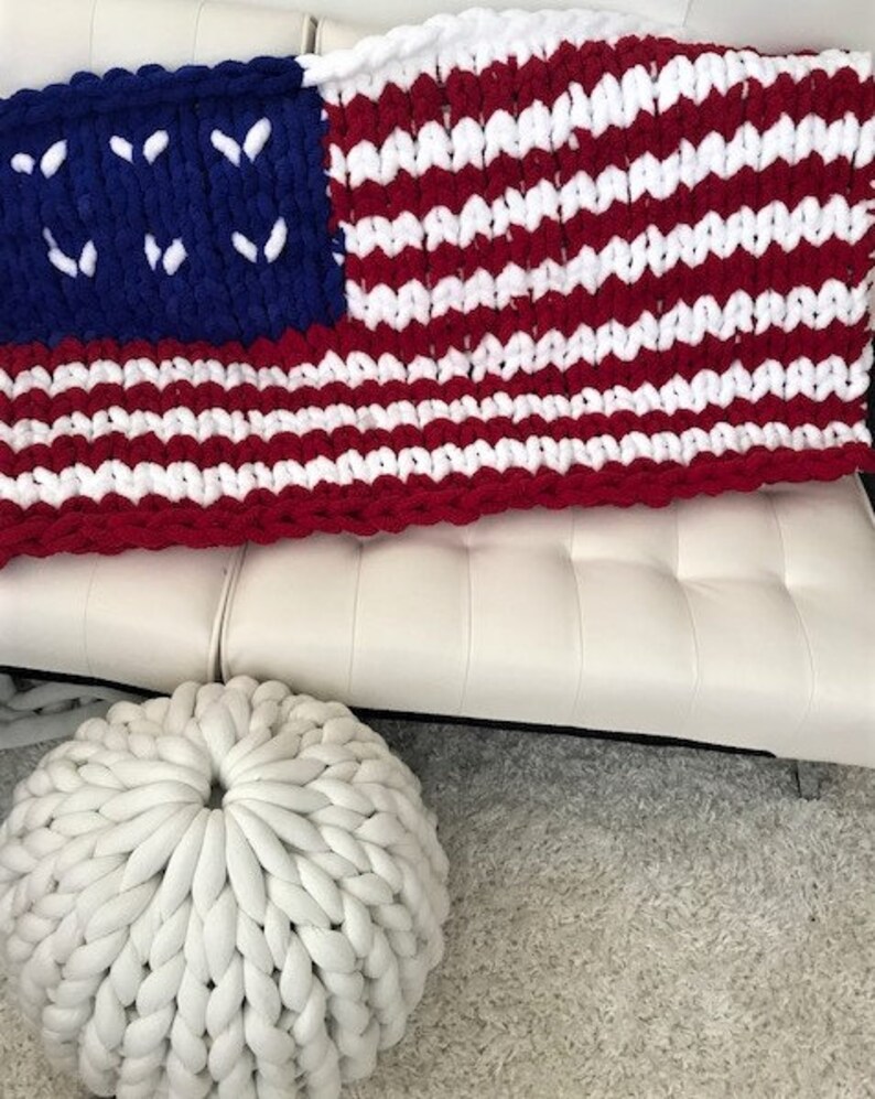 K&ouml;nnte beinhalten: Eine grob gestrickte Decke im Stil der amerikanischen Flagge mit roten und wei&szlig;en Streifen und einem blauen Feld mit wei&szlig;en Sternen. Ein wei&szlig;er, runder, gestrickter Pouf befindet sich im Vordergrund. Die Decke ist &uuml;ber ein wei&szlig;es Sofa drapiert.