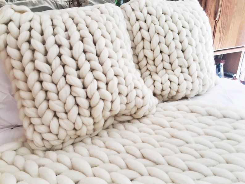 May include: Two white chunky knit throw pillows on a white chunky knit throw blanket. The pillows and blanket are made of thick, soft yarn.