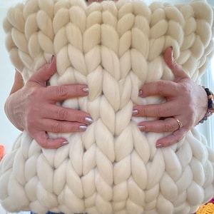 May include: A large, white, chunky knit pillow with a cable knit pattern. The pillow is being held by a person's hands.