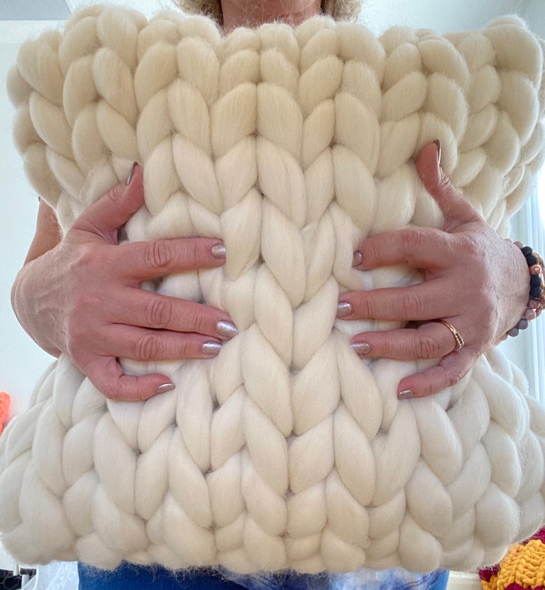 May include: A large, white, chunky knit pillow with a cable knit pattern. The pillow is being held by a person's hands.