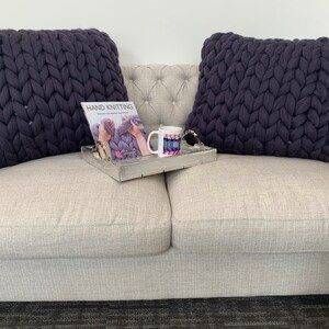 May include: Two dark gray chunky knit throw pillows on a light gray couch with a book titled "Hand Knitting" and a mug with a colorful design on a wooden tray.