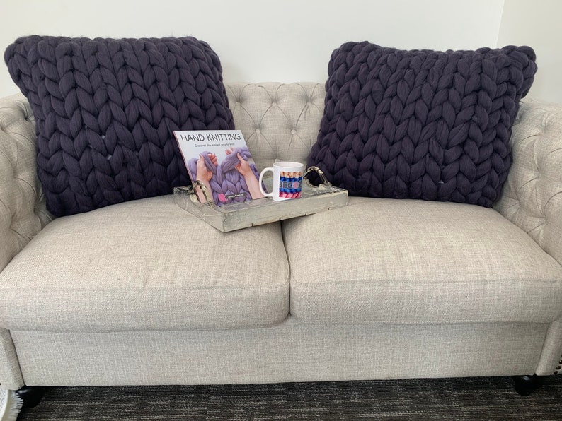 May include: Two dark gray chunky knit throw pillows on a light gray couch with a book titled "Hand Knitting" and a mug with a colorful design on a wooden tray.