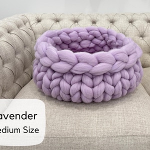 May include: A round, chunky knit basket in a soft lavender color, featuring thick, braided yarn loops. A white rectangular label on the lower left corner displays the black text Lavender Medium Size in a clean, sans-serif font.