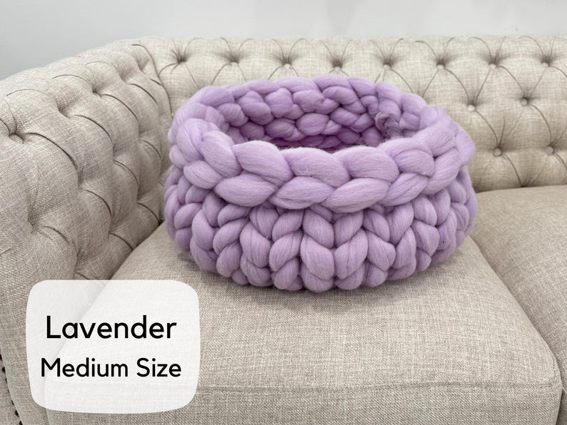 May include: A round, chunky knit basket in a soft lavender color, featuring thick, braided yarn loops. A white rectangular label on the lower left corner displays the black text Lavender Medium Size in a clean, sans-serif font.