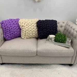 May include: Three chunky knit throw pillows in purple, white, and navy blue on a light gray couch.
