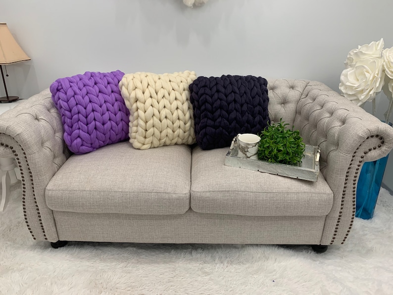 May include: Three chunky knit throw pillows in purple, white, and navy blue on a light gray couch.