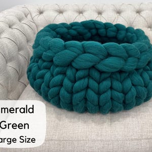 May include: A round, chunky knit pet bed made of thick, emerald green yarn. The circular structure features a braided texture with a deep, open center. A white rectangular label in the corner displays the text Emerald Green Large Size.