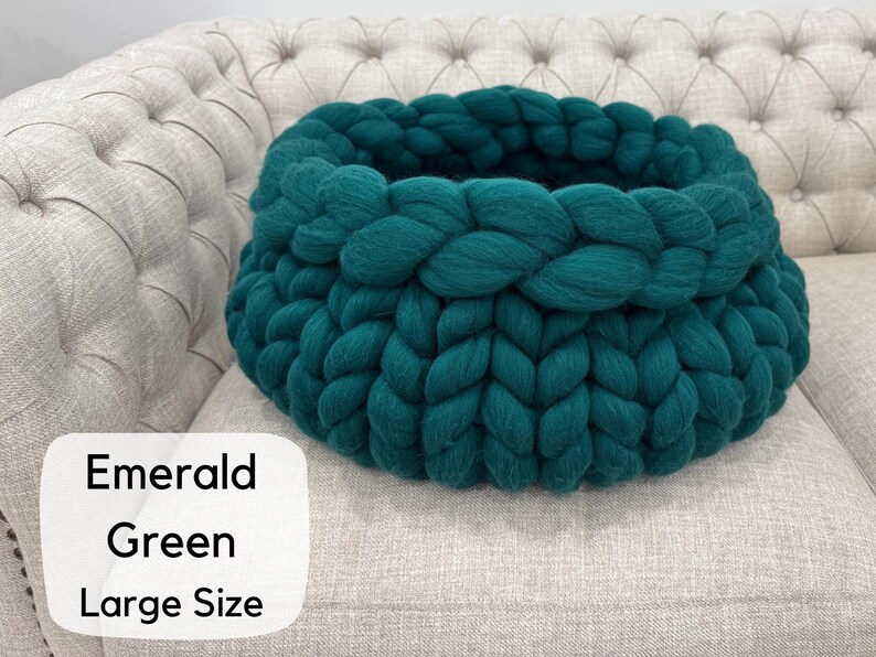 May include: A round, chunky knit pet bed made of thick, emerald green yarn. The circular structure features a braided texture with a deep, open center. A white rectangular label in the corner displays the text Emerald Green Large Size.