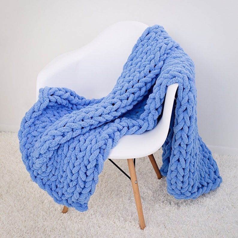 May include: A light blue chunky knit throw blanket draped over a white chair. The blanket is made with a thick yarn and has a textured, cable knit pattern.