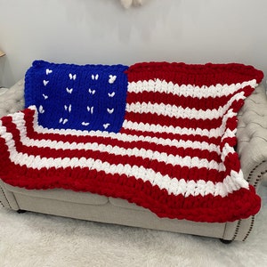 K&ouml;nnte beinhalten: Eine grob gestrickte Decke, die &uuml;ber einem Sofa drapiert ist und das Design der amerikanischen Flagge aufweist. Die Decke hat rote und wei&szlig;e Streifen mit einem blauen Quadrat in der Ecke, das mit wei&szlig;en Sternen verziert ist.