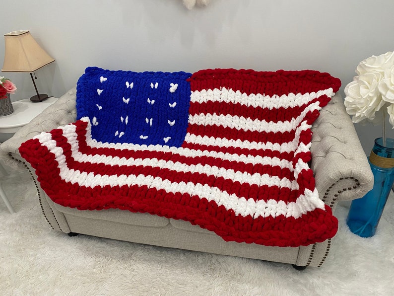 K&ouml;nnte beinhalten: Eine grob gestrickte Decke, die &uuml;ber einem Sofa drapiert ist und das Design der amerikanischen Flagge aufweist. Die Decke hat rote und wei&szlig;e Streifen mit einem blauen Quadrat in der Ecke, das mit wei&szlig;en Sternen verziert ist.