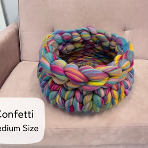 May include: A round, chunky knit basket crafted from thick, multicolored yarn in shades of pink, blue, yellow, and teal. The basket features a braided texture and includes a white label with the text Confetti Medium Size.