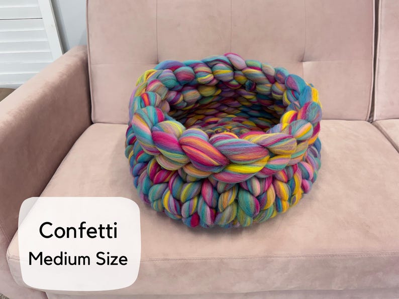 May include: A round, chunky knit basket crafted from thick, multicolored yarn in shades of pink, blue, yellow, and teal. The basket features a braided texture and includes a white label with the text Confetti Medium Size.