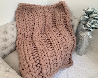 May include: A chunky knit throw blanket in a soft, dusty rose color. The blanket is made of thick yarn and has a cable knit pattern. It is draped over a light gray couch with a white fluffy rug underneath.