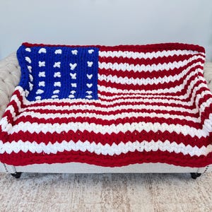 K&ouml;nnte beinhalten: Eine grob gestrickte Decke, die &uuml;ber einem Sofa drapiert ist und das Design der amerikanischen Flagge zeigt. Die Decke hat rote und wei&szlig;e Streifen mit einem blauen Quadrat in der Ecke, das mit wei&szlig;en Sternen verziert ist. Die Decke ist aus dickem Garn gefertigt, ca. 150 cm x 200 cm.