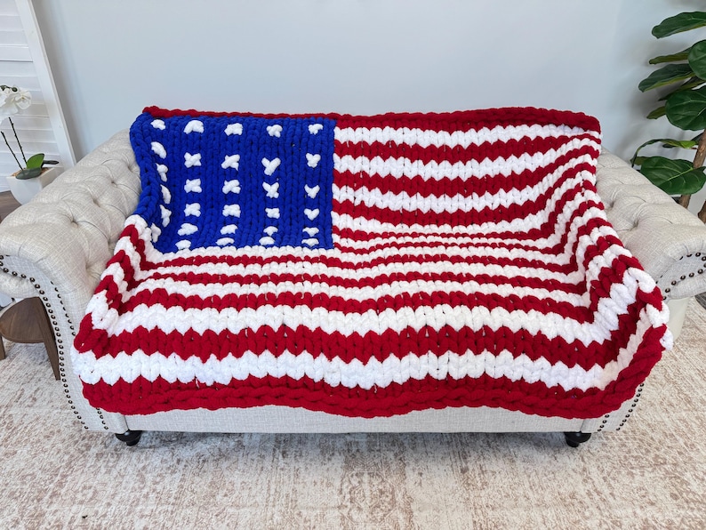 K&ouml;nnte beinhalten: Eine grob gestrickte Decke, die &uuml;ber einem Sofa drapiert ist und das Design der amerikanischen Flagge zeigt. Die Decke hat rote und wei&szlig;e Streifen mit einem blauen Quadrat in der Ecke, das mit wei&szlig;en Sternen verziert ist. Die Decke ist aus dickem Garn gefertigt, ca. 150 cm x 200 cm.