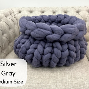 May include: A medium size basket crafted from thick, chunky silver gray yarn in a braided knit pattern. The basket features a rounded shape with a wide opening and a textured, soft appearance created by the large, interlocking yarn loops.