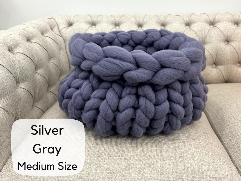 May include: A medium size basket crafted from thick, chunky silver gray yarn in a braided knit pattern. The basket features a rounded shape with a wide opening and a textured, soft appearance created by the large, interlocking yarn loops.