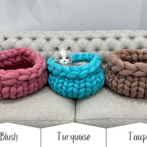 May include: Three round, chunky knit pet beds in blush, turquoise, and taupe. The turquoise bed contains a small, fluffy, grey and white plush cat toy. Each bed features a thick, braided yarn construction with a deep, circular opening.