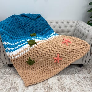 Peut inclure: Une couverture en tricot épais sur le thème de la plage. La couverture présente un océan bleu, des vagues blanches et du sable beige. Des accents de tortue verte et d'étoile de mer corail sont cousus sur la couverture. La couverture est drapée sur un canapé beige.