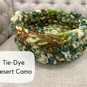 Puede incluir: Una cama redonda para mascotas tejida a mano con un patr&oacute;n de camuflaje des&eacute;rtico te&ntilde;ido anudado. La cama presenta una mezcla de hilo verde, marr&oacute;n y crema. El texto "Tie-Dye Desert Camo" se muestra en una etiqueta blanca.