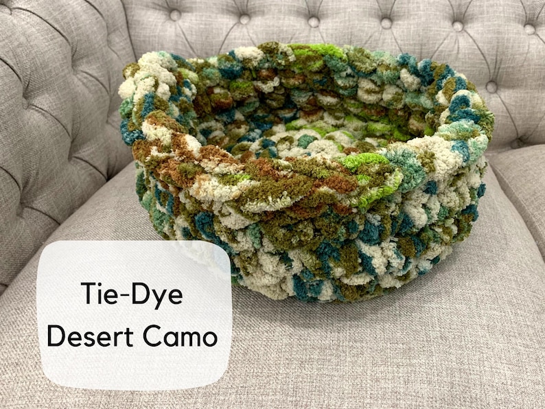 Puede incluir: Una cama redonda para mascotas tejida a mano con un patr&oacute;n de camuflaje des&eacute;rtico te&ntilde;ido anudado. La cama presenta una mezcla de hilo verde, marr&oacute;n y crema. El texto "Tie-Dye Desert Camo" se muestra en una etiqueta blanca.