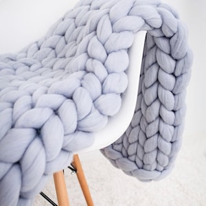 May include: A chunky knit blanket in a soft light gray color, featuring large, thick yarn loops woven into a dense, braided pattern. The heavy, textured textile is draped over the back and seat of a white chair with light wooden legs.