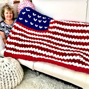 K&ouml;nnte beinhalten: Eine gro&szlig;e, grob gestrickte Decke im Stil der amerikanischen Flagge. Die Decke hat ein blaues Feld mit wei&szlig;en Sternen und rot-wei&szlig;en Streifen. Die Decke ist &uuml;ber ein wei&szlig;es Sofa drapiert. Ma&szlig;e in Zentimetern.