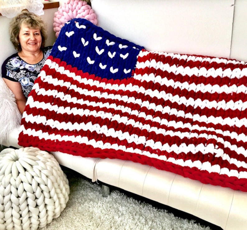 K&ouml;nnte beinhalten: Eine gro&szlig;e, grob gestrickte Decke im Stil der amerikanischen Flagge. Die Decke hat ein blaues Feld mit wei&szlig;en Sternen und rot-wei&szlig;en Streifen. Die Decke ist &uuml;ber ein wei&szlig;es Sofa drapiert. Ma&szlig;e in Zentimetern.